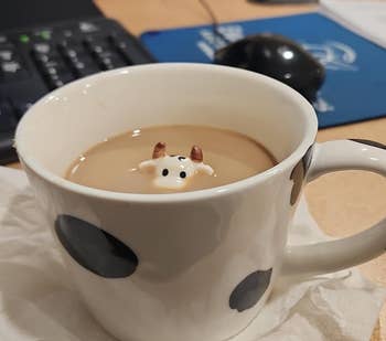 Cow-themed mug with a ceramic cow figure peeking out from the coffee inside, placed on a napkin near a computer keyboard and mouse