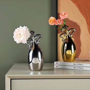 Two ornate vases, one silver and one gold, with flowers, displayed on a table against a painting backdrop