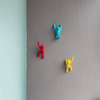 Three wall hooks shaped like climbers in red, blue, and yellow adorn a patio wall, with a leafy plant in the corner