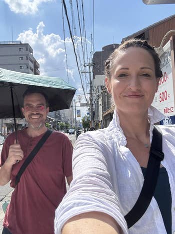 Two people smile while walking on a city street, one holding an umbrella. Urban background with buildings and power lines. Casual attire