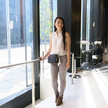 Person in a sleeveless blouse, gray pants, and ankle boots, standing by a window in a modern store