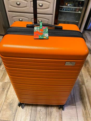 Orange hard-shell suitcase with a floral luggage tag stands on a wooden floor next to a kitchen cabinet