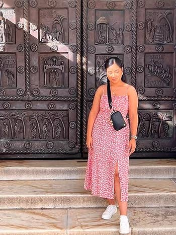 Person in a patterned sundress with a crossbody bag and sneakers stands on steps before an ornate metal door