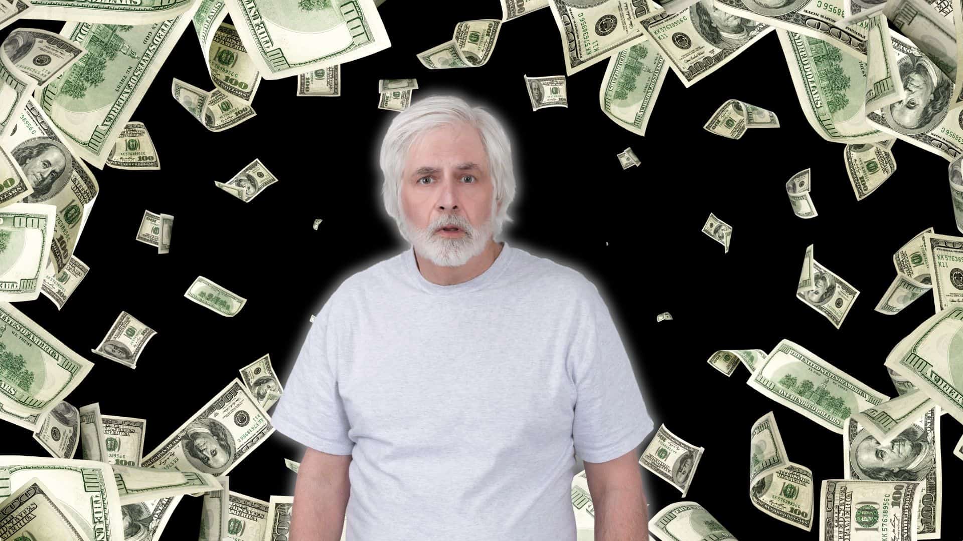 A light-skinned older man with white hair and a beard, wearing a light grey t-shirt, looks directly forward with a wide-eyed, slightly surprised expression. He is centered against a black background, surrounded by numerous green and white $100 bills falling and floating around him, creating a vortex-like effect of money.
