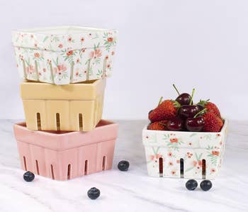 Stacked ceramic baskets with floral designs and one filled with strawberries and cherries on a marble surface, alongside scattered blueberries