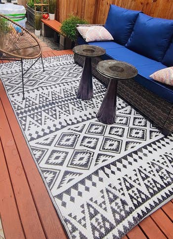 Outdoor patio with geometric-patterned rug, wicker sofa with plush cushions, wooden fencing, and small tables. Cozy setup for relaxing or entertaining