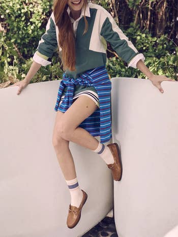 Person sitting on a wall wearing a casual preppy outfit with a collared top, striped shorts, and loafers