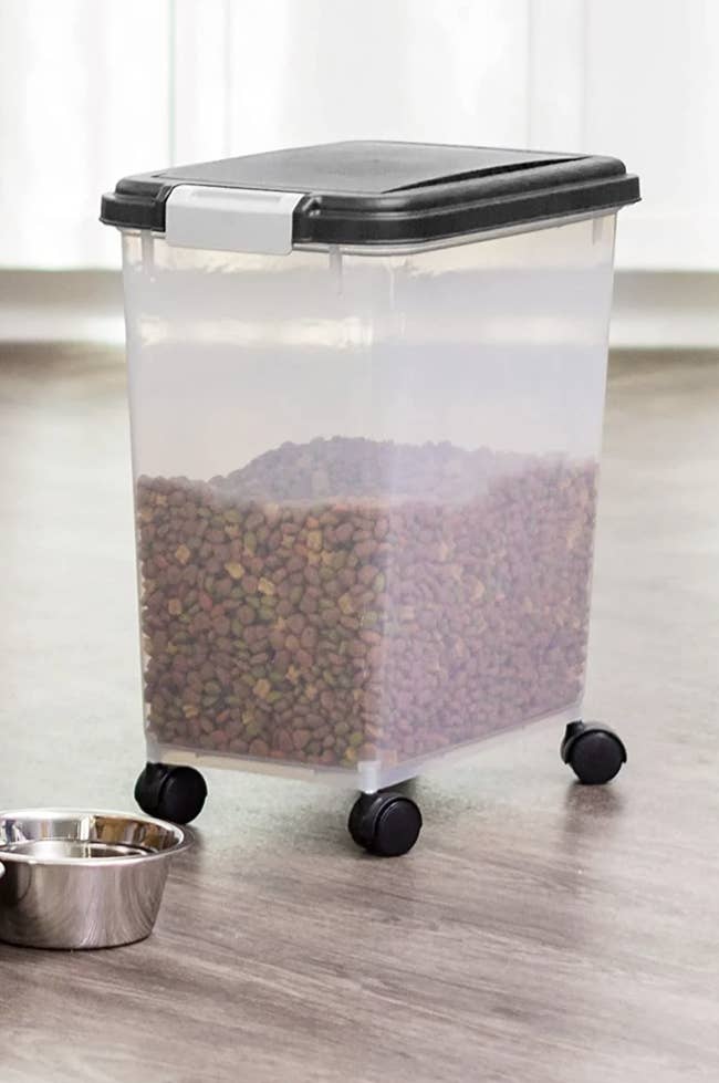 Large, clear plastic container on wheels filled with dry pet food. Metal bowl nearby on the floor