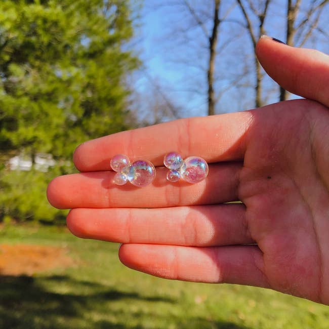A hand holding two small, clear bubble clusters outdoors, with trees and grass in the background