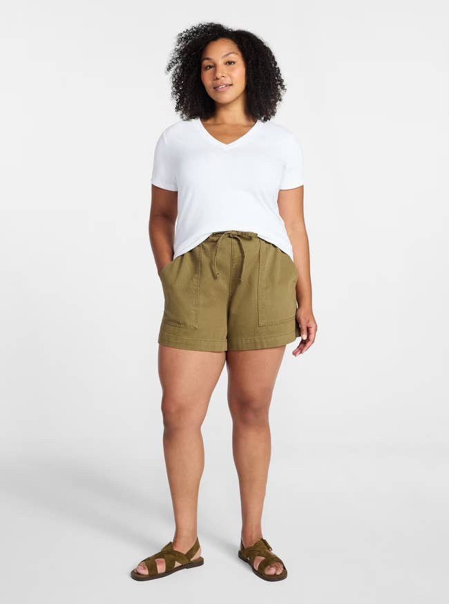 Person wearing a casual white V-neck T-shirt, olive shorts, and sandals, standing and smiling against a plain background