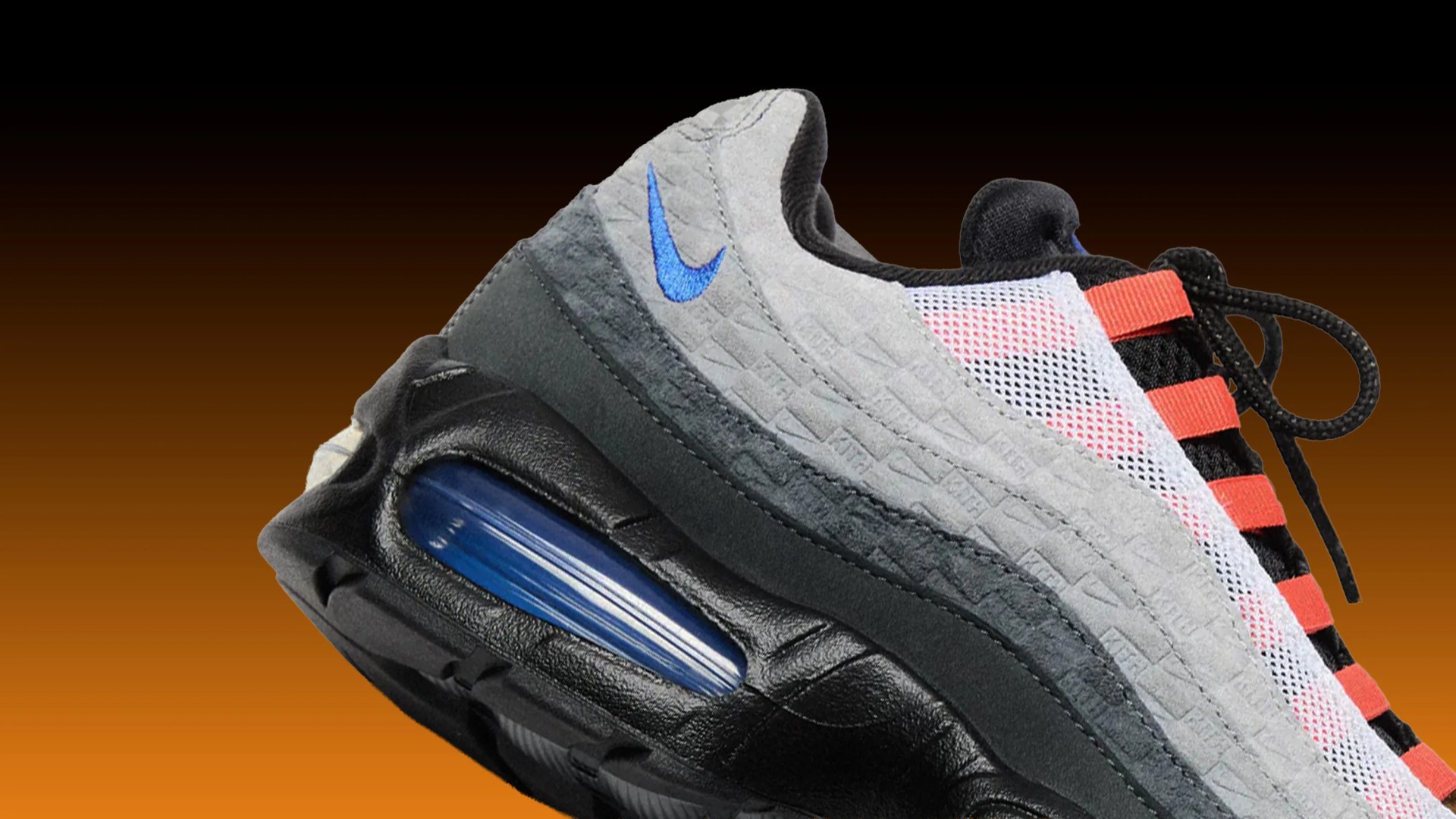 Close-up of a Nike sneaker with grey suede, black leather sole, blue air cushion, white mesh, and orange lace loops.