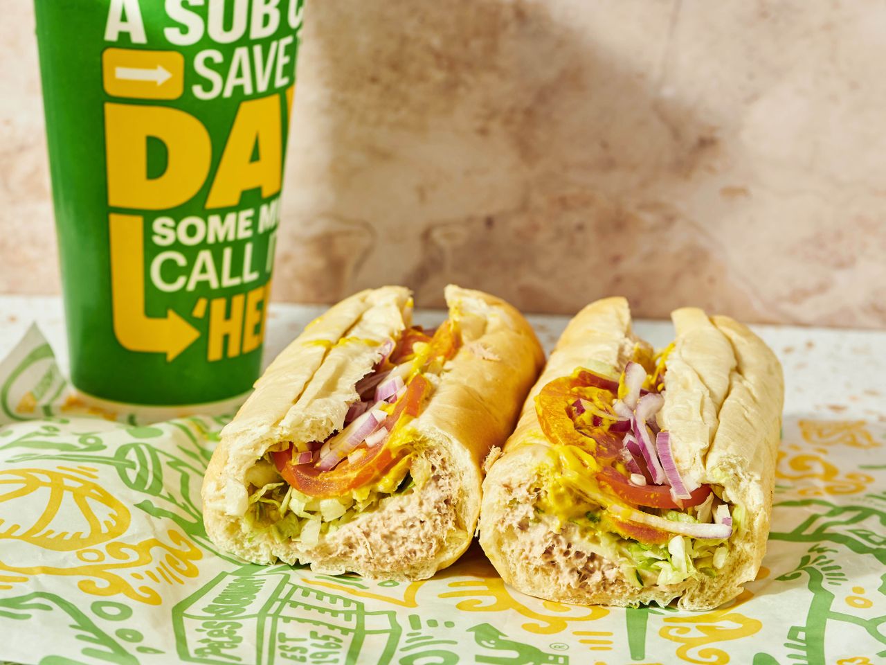 Subway tuna sandwich with lettuce, tomatoes, and red onion next to a green drink cup.