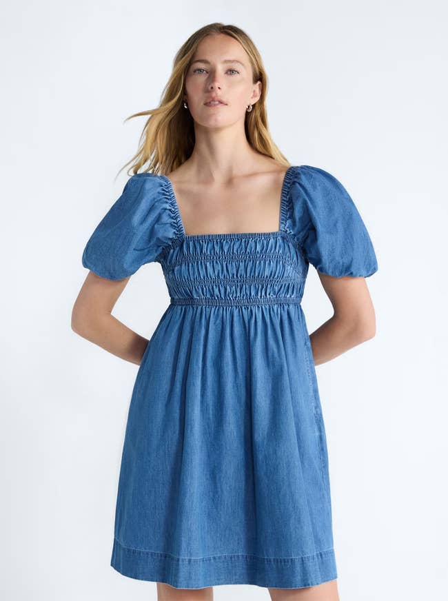 Person in a denim dress with puffed sleeves and a shirred bodice poses with hands on hips