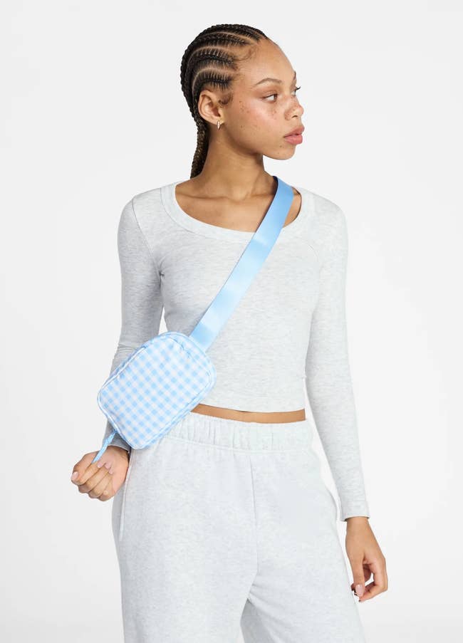 Person wearing a light gray top and sweatpants, modeling a blue checkered crossbody bag on a white background