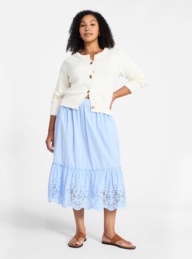 Person wearing a buttoned cardigan, blue eyelet midi skirt, and sandals, standing with a relaxed pose