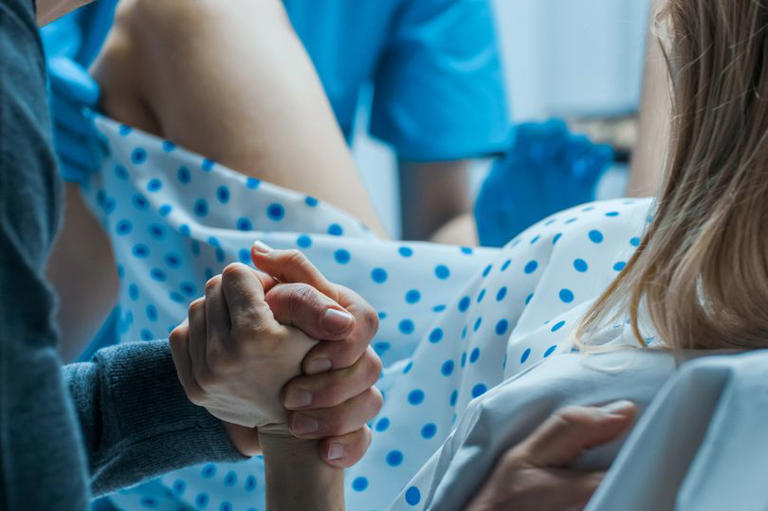 Emergency In the Hospital: Woman Giving Birth, Husband Holds Her Hand in Support, Obstetricians Assisting. Modern Delivery Ward with Professional Midwives.