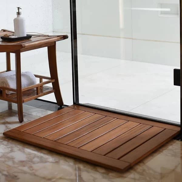 Bathroom scene with a wooden bathmat, a small table with a towel and soap dispenser next to a glass shower door, on a marble floor