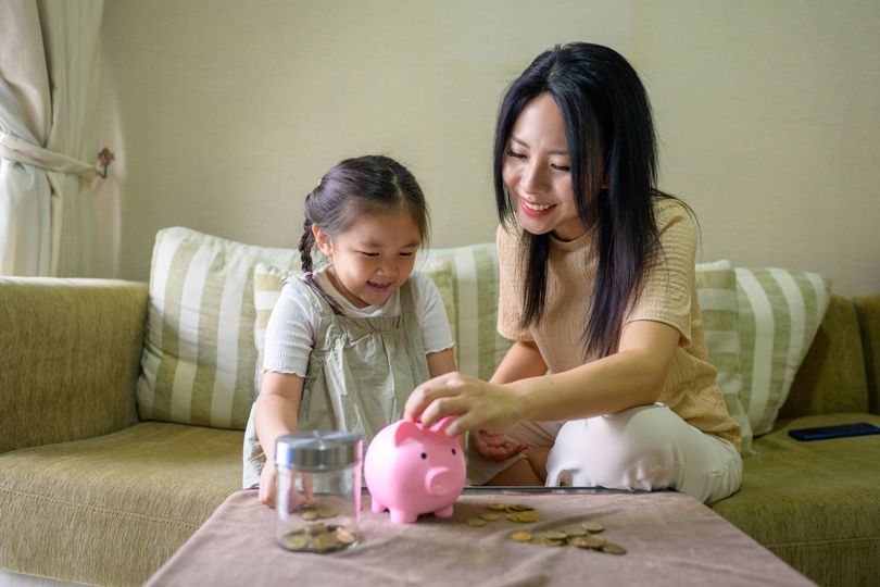 A young Asian woman and her daughter are happily saving coins in a pink piggy bank on a table at home.