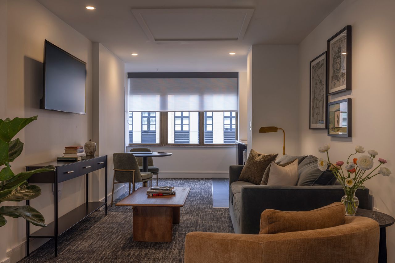 A cozy hotel room with seating furniture a coffee table a television on the wall and a view of windows in the background