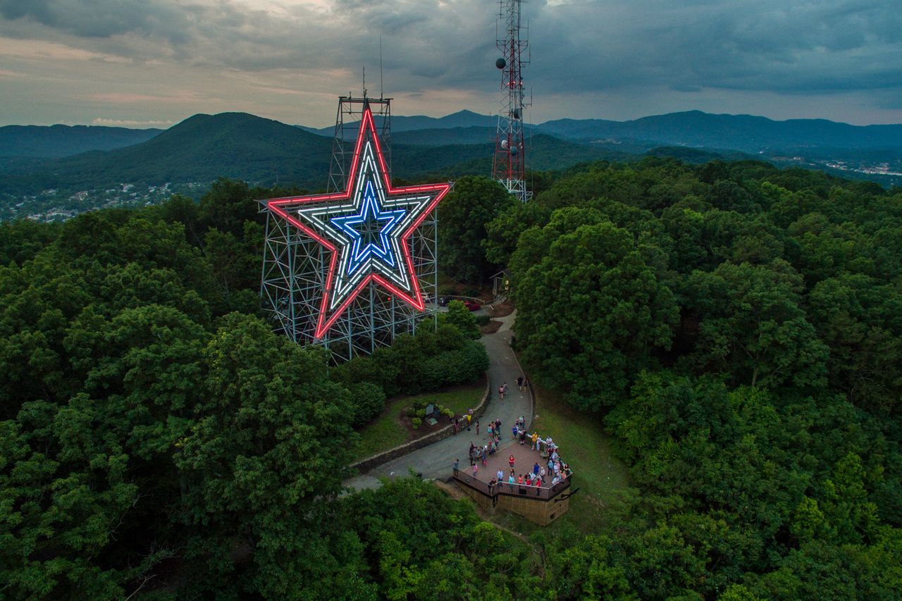 Large illuminated star structure on a hill surrounded by forested landscape and visitors gathered at its base