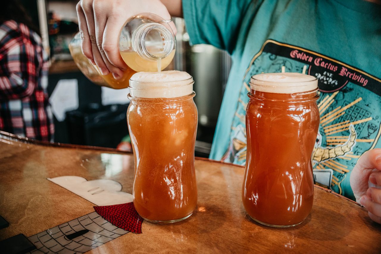Two glasses of beer being poured at a bar with a person wearing a graphic shirt in the background