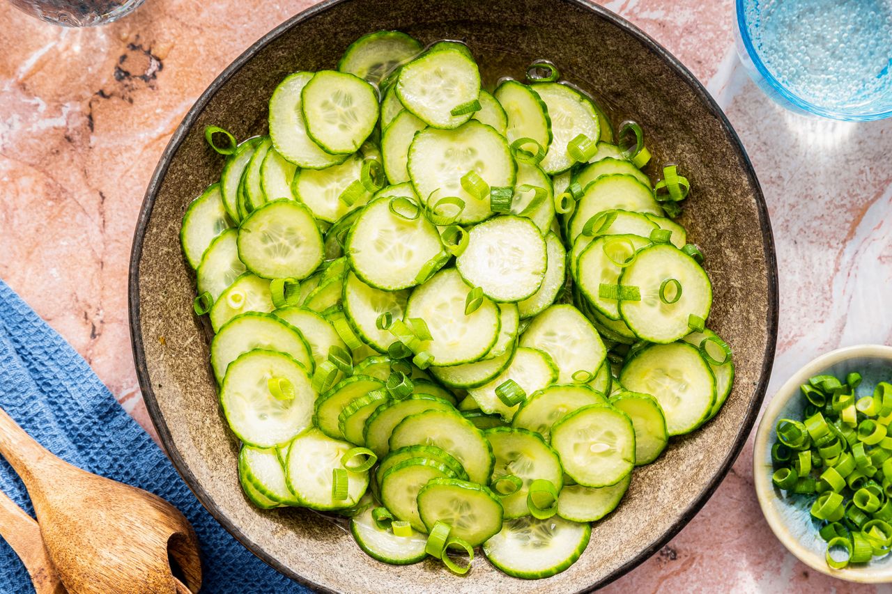 cucumber salad in a bowl at a table setting with a bowl of slcied scallions, serving utensils on a table napkin, and a glass of water