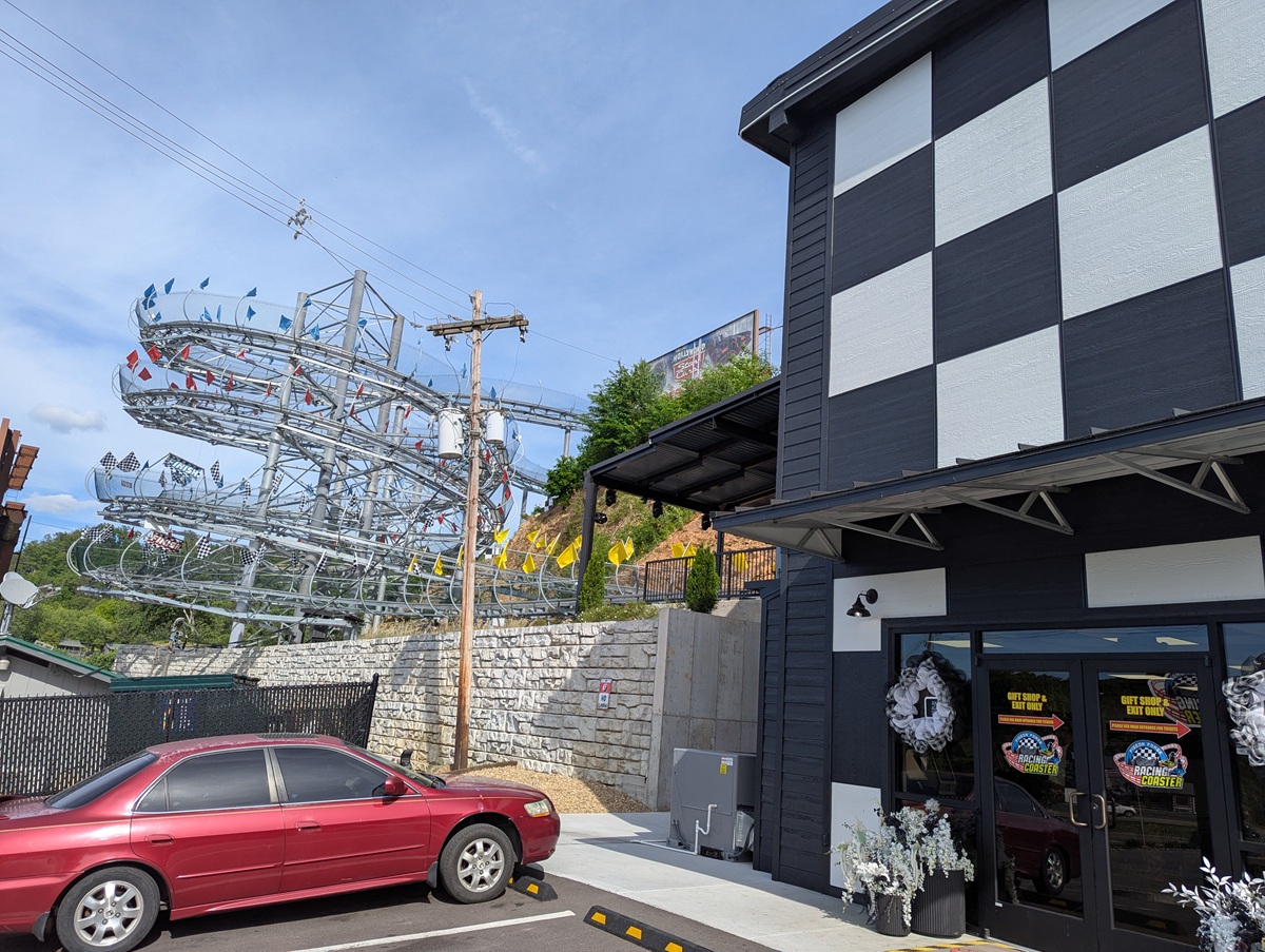 The Pigeon Forge Racing Coaster in Pigeon Forge, Tennessee, a metal alpine coaster track with twists in tight loops up a hillside beside a black and white striped building, with colorful flags lining the rails. The red car in the parking lot and the coaster’s exposed steel curves make the scene feel lively and amusement park adjacent.
