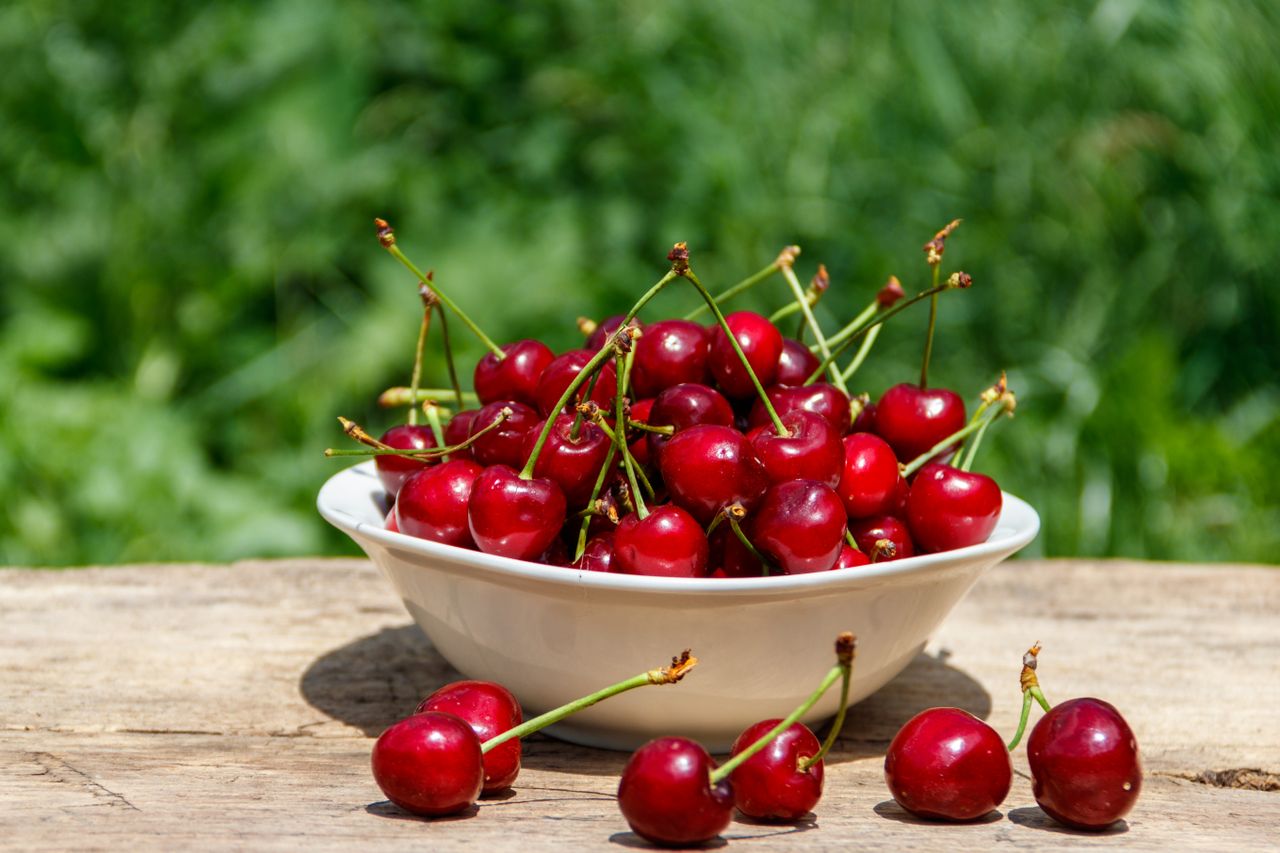 A bowl filled with cherries placed on a wooden surface outdoors.