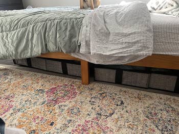 Under-bed storage baskets are neatly arranged under a bed with a colorful floral rug nearby