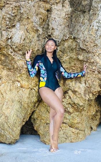 Person wearing a floral long-sleeve swimsuit poses against a rocky background, suggesting a beach setting for a shopping or style article