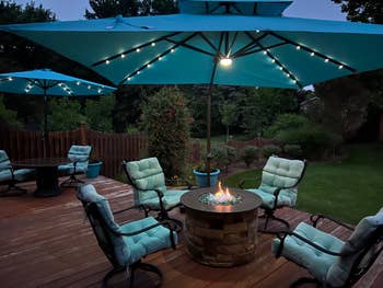 Cozy outdoor patio with cushioned chairs, fire pit, and large umbrellas with lights, set in a lush garden. Ideal for evening gatherings