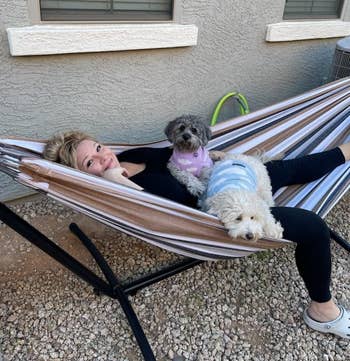 Person relaxing on a hammock with two small dogs in sweaters, outdoors in a yard with a window and gravel ground