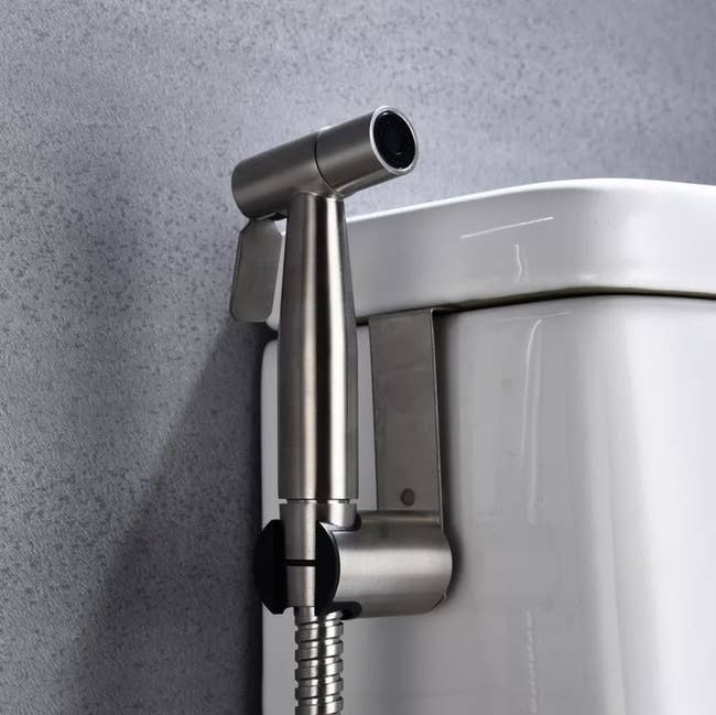 A stainless steel handheld bidet sprayer is mounted on the side of a toilet tank in a modern bathroom setting
