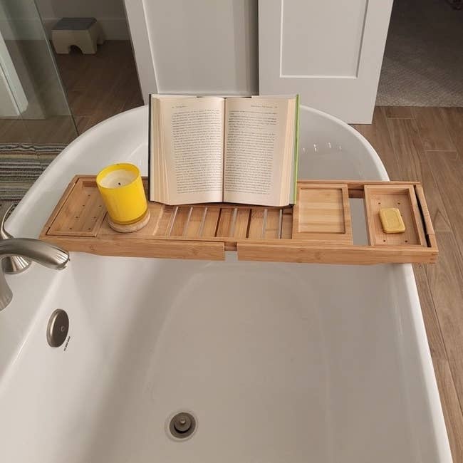 A bathtub caddy with a yellow candle, book, and soap rests across a bathtub, suggesting a relaxing bath experience