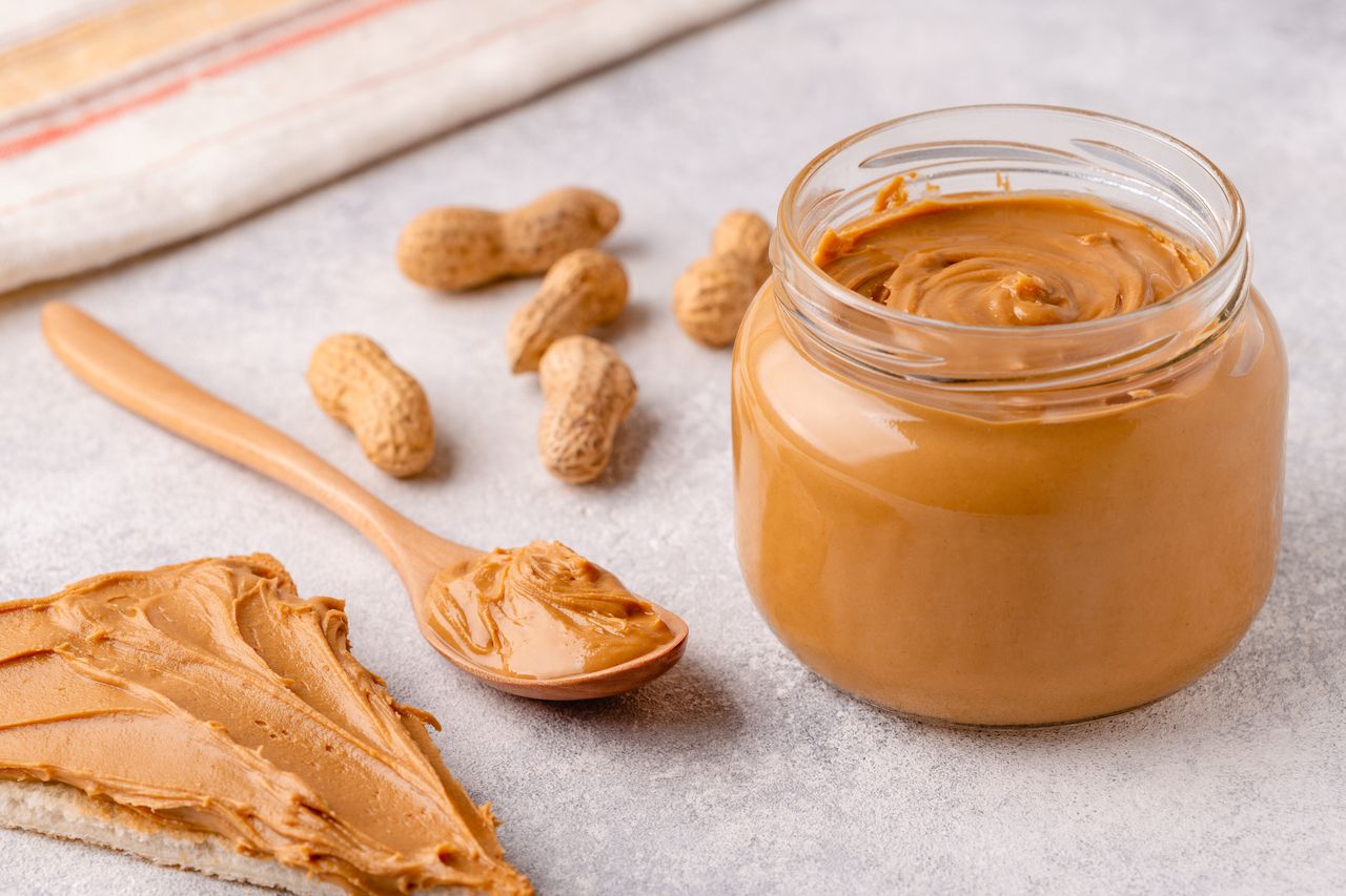 A jar of peanut butter with a spoon and peanuts toast spread with peanut butter beside it