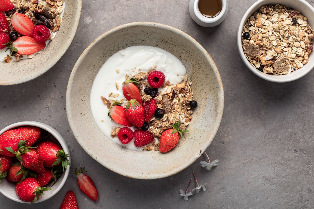 A bowl of yogurt topped with granola strawberries and raspberries surrounded by bowls of strawberries granola and a small cup of syrup