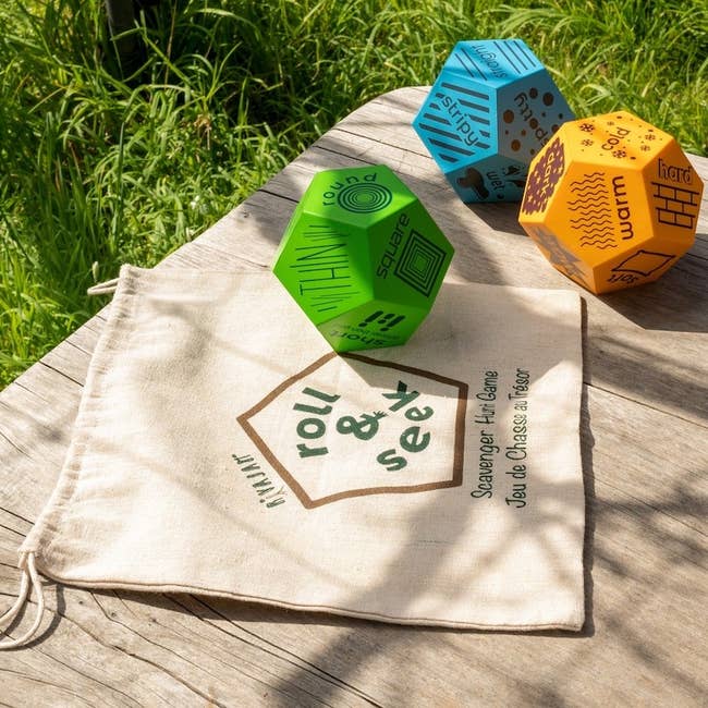Three geometric dice with activity prompts on a wooden table, next to a cloth bag labeled