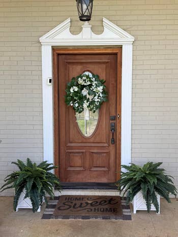 Wooden front door with a glass panel, adorned with a floral wreath, flanked by green potted ferns, and a