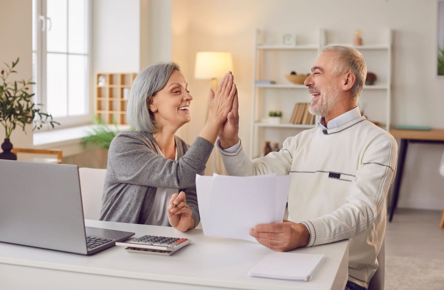 A smiling senior couple high-fives at a white desk in a brightly lit room. The woman, with gray hair, wears a gray cardigan. The man, with gray hair and a beard, wears a white sweater. On the desk are a silver laptop, a calculator, several white papers, and a small notebook. A green houseplant and a window are visible in the background.