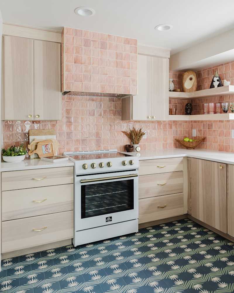 Modern kitchen featuring light wood cabinets, pink tiled backsplash, white stove, and patterned blue-green floor tiles.