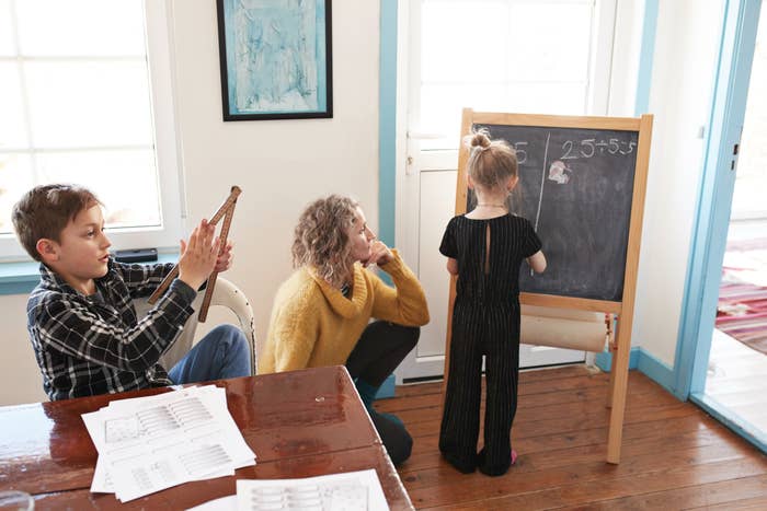 Child writing on chalkboard while adult and another child watch, with printed papers on nearby table