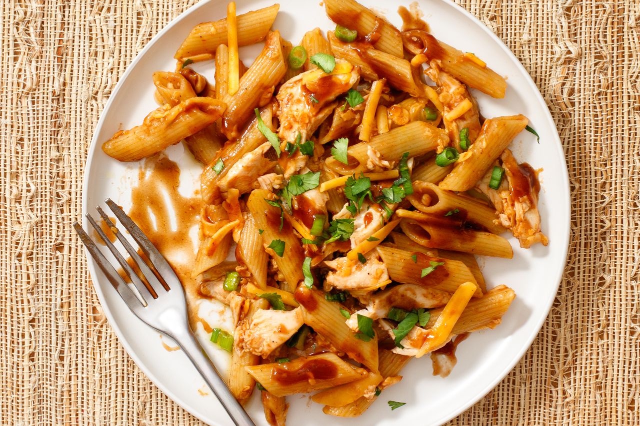 A plate of pasta with barbecue chicken garnished with herbs and sauce and a fork beside it on a textured surface