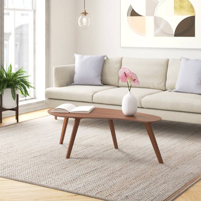 Modern living room with a beige sofa, wooden coffee table, and decorative vase. Minimalist wall art and a potted plant enhance the cozy ambiance