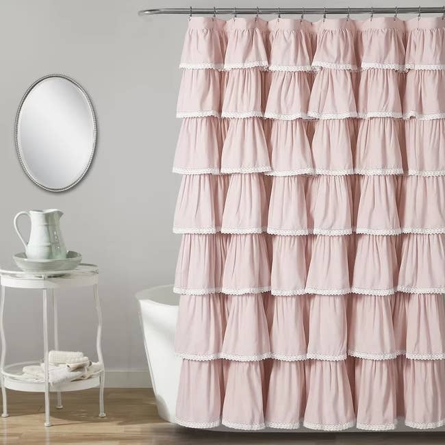 Frilly tiered shower curtain with lace trim hung in a bathroom; includes round mirror and small table with porcelain decor