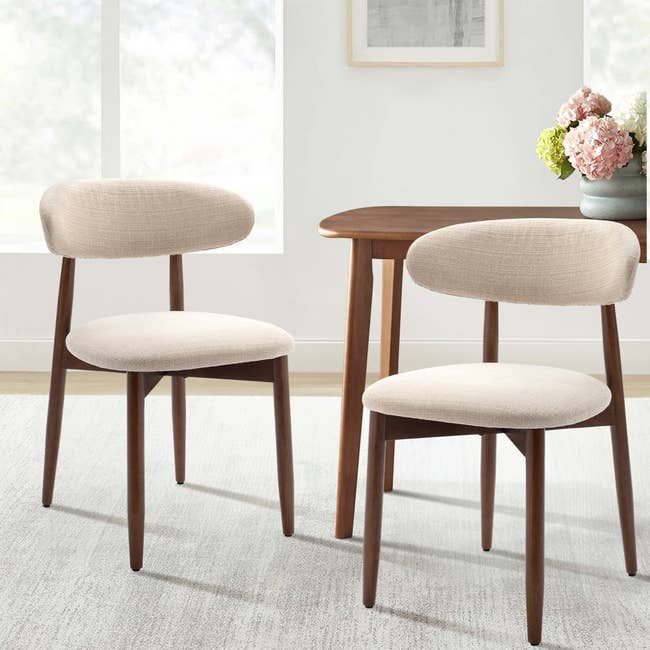 Two modern dining chairs with curved backs and light fabric upholstery, next to a wooden table, set in a bright, minimalist room