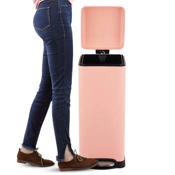 Person in jeans demonstrating a pink smart trash can with an open lid and foot pedal, highlighting its styling for a shopping feature
