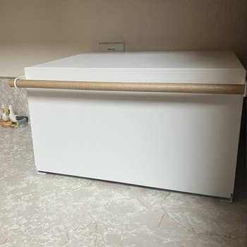 Modern white bread box with a wooden handle sitting on a kitchen countertop under cabinets