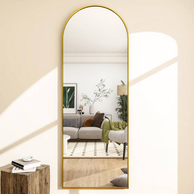 Arch-shaped gold mirror reflecting a modern living room with a couch, green accents, plants, and geometric patterns; wooden stool in foreground