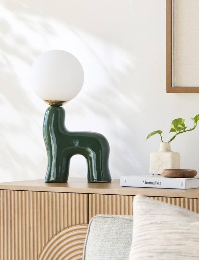Modern table lamp with a white globe on a unique, green ceramic base on a wooden cabinet next to a minimalist book and a small plant