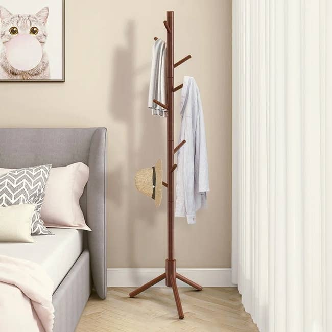 Bedroom scene with a wooden coat rack holding a hat and clothes next to a bed, featuring a cat artwork on the wall
